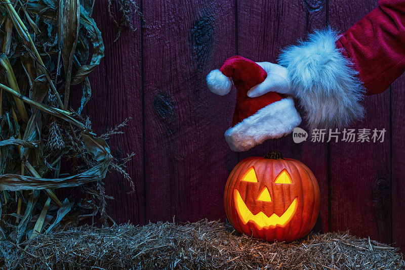 圣诞老人把圣诞帽放在一个快乐雕刻的杰克O' Lantern上，在干草和干玉米杆